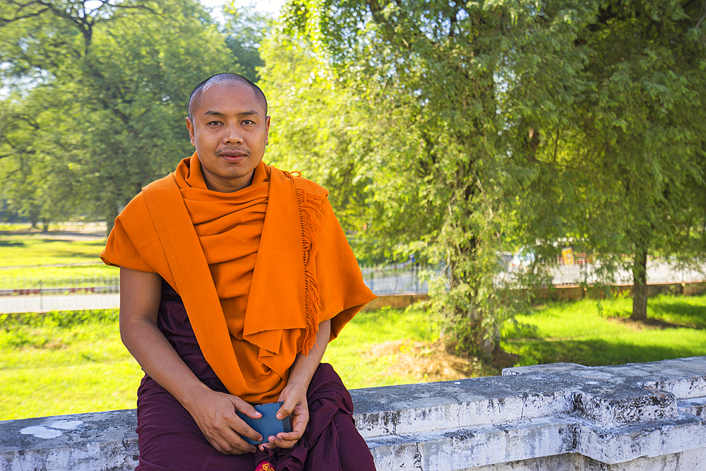 Monk in saffron robe sitting on wall with his cell phone in hand, Royal Palace, Mandalay, Myanmar (Burma), Asia