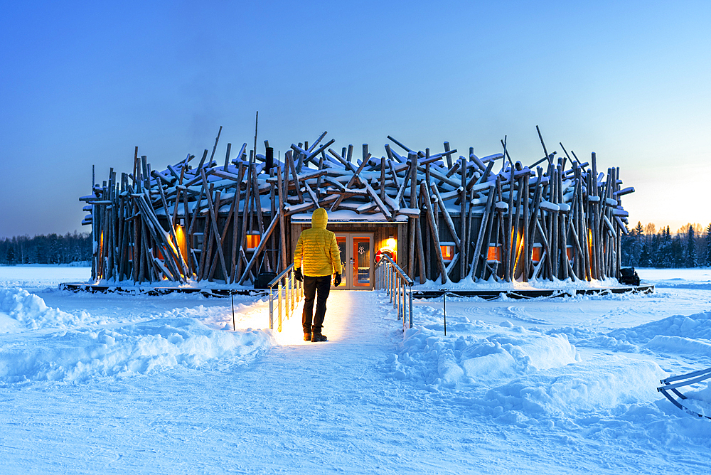 Person stands on the bridge connecting the main building of the illuminated Arctic Bath hotel made of logs, dusk time, Harads, Swedish Lapland, Norrbotten, Sweden, Scandinavia, Europe