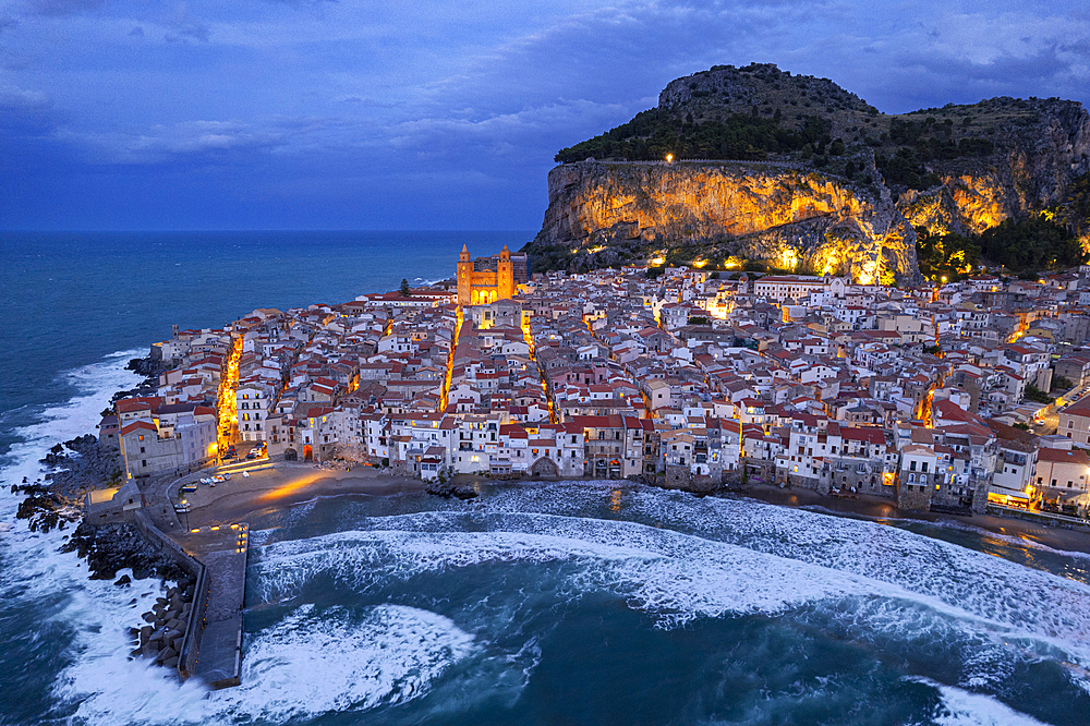 The illuminated fishing village of Cefalu with the rock on top of the old town seen from the drone at dusk, Cefalu, Palermo province, Tyrrhenian Sea, Sicily, Italy, Mediterranean, Europe