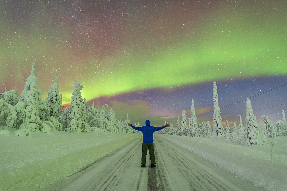 Rear view of man with outstratched arms in the middle of an empty slippery road crossing the Arctic forest while admiring the Northern Lights (Aurora Borealis), Finnish Lapland, Finland, Europe