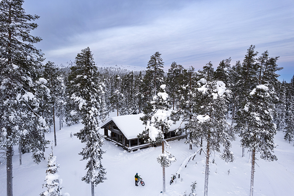 Drone view of a cyclist with fat bike standing in the middle of the tall-tree coniferous forest covered with snow in front of a mountain hut, Finnish Lapland, Finland, Scandinavia, Europe