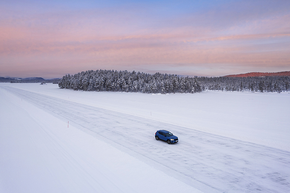 Aerial view of a car traveling on icy road on top of a frozen lake surrounded by coniferous forest covered with snow, a dusk, Jokkmokk, Norrbotten, Swedish Lapland, Sweden, Scandinavia, Europe