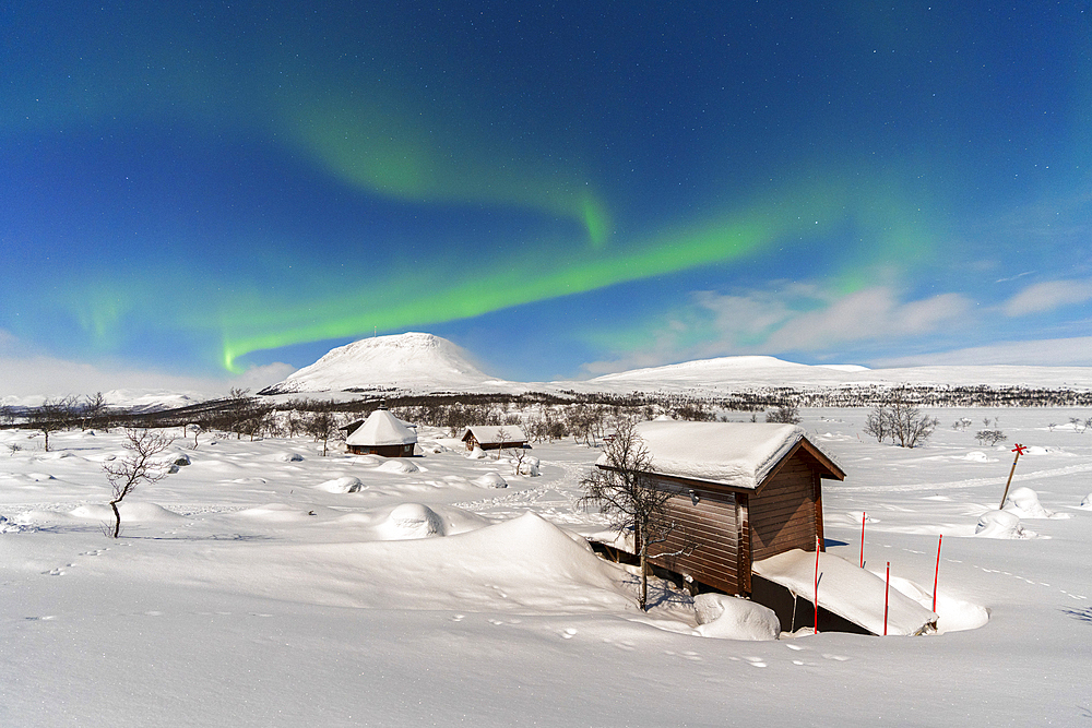 Green Northern Lights (Aurora Borealis) above typical wooden huts in the snow-covered landscape lit by full moon, Kilpisjarvi, Enontekio municipality, Finnish Lapland, Finland, Scandinavia, Europe