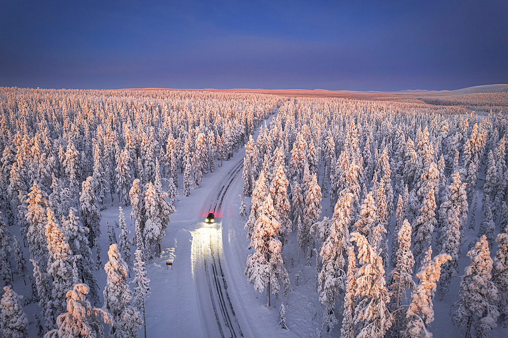 Aerial view of car on icy road and illuminated headlamps driving in the snowcapped forest, at dawn, Akaslompolo, Kolari, Pallas-Yllastunturi National Park, Lapland region, Finland, Europe