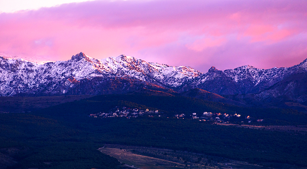 Dramatic purple sunset above the snowy rugged summits of los cumbres verdes. A part of the Sierra Nevada with a little town on the foot of the mountain range. View from Granada, Spain.