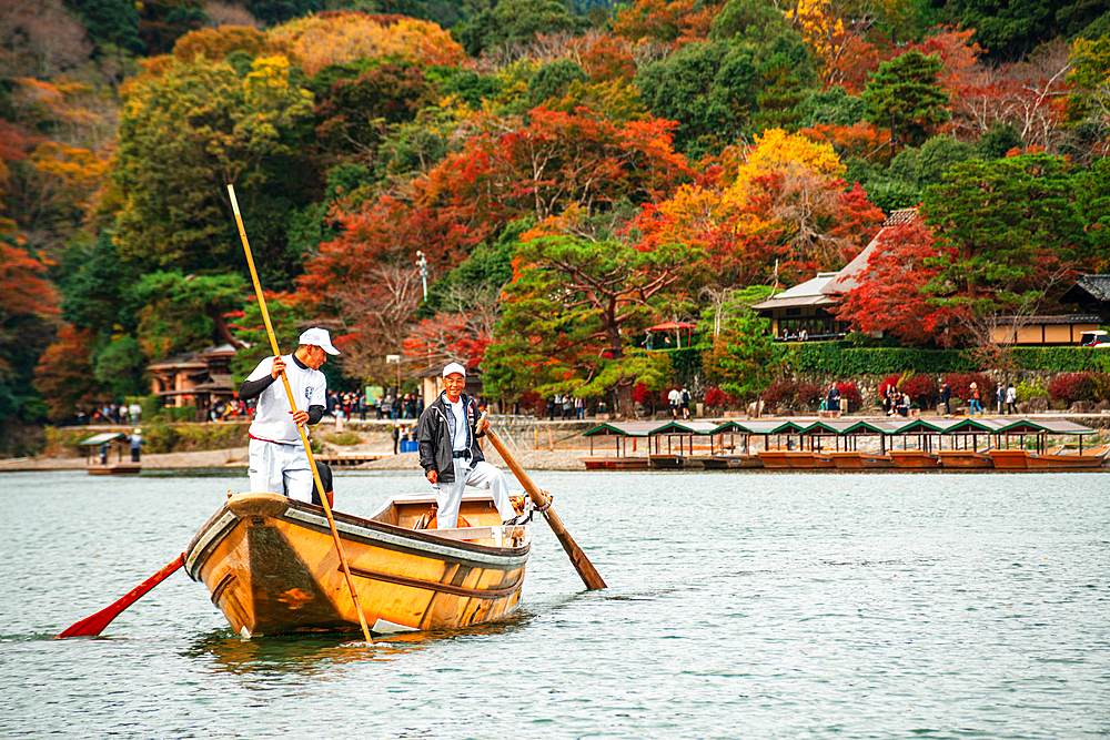 A traditional wooden boat navigates the Katsura River in Arashiyama, Kyoto, with vibrant autumn foliage in the background.