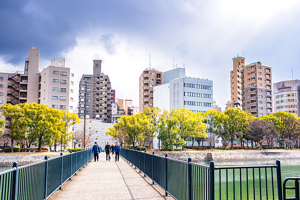 Pedestrian walkway over the river in Hiroshima leading to a cityscape of mid-rise buildings and trees under dramatic clouds