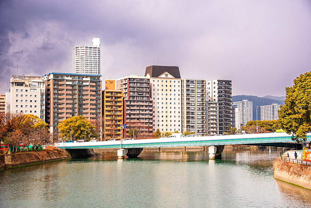 Colorful apartment buildings along the riverside in Hiroshima with a bridge crossing the calm water under dramatic cloudy skies