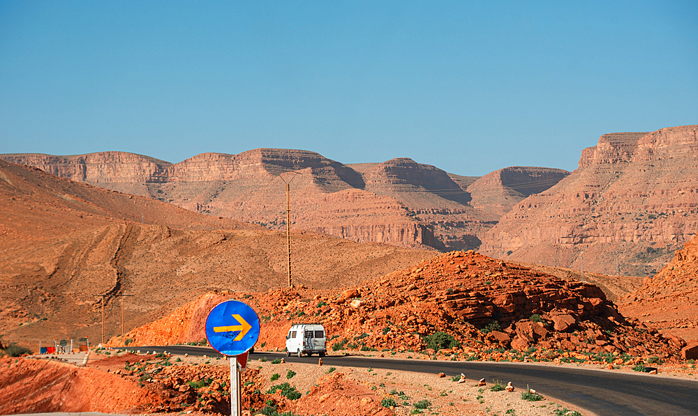 White van navigating a sharp desert road bend in Parc National Al Hassan Addakhil, red rocks and distant mountains under a clear blue sky