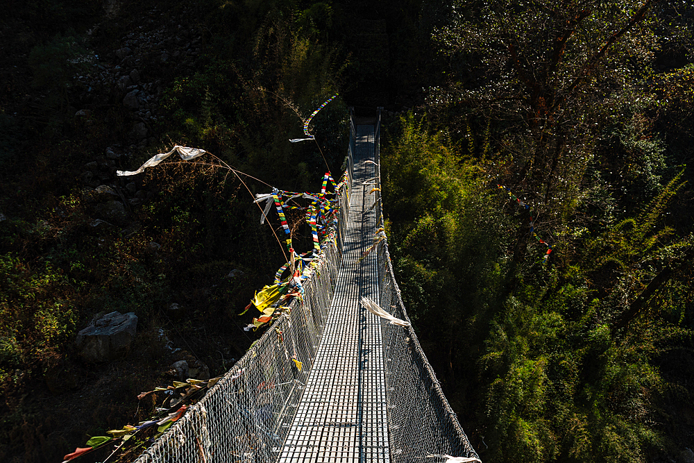 Narrow suspension bridge with fluttering prayer flags crossing deep forest gorge in Langtang Valley, Nepal, seen from a moody elevated angle