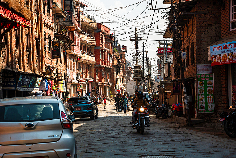 Motorcyclists and pedestrians navigating a sunlit narrow street lined with traditional and modern buildings in Bhaktapur