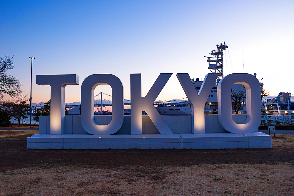 Large illuminated TOKYO sign at sunset with Rainbow Bridge and city skyline in the background at Odaiba Seaside Park, Tokyo, Japan