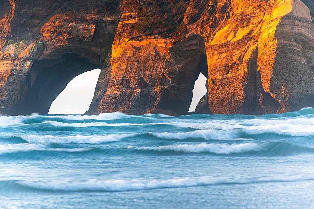 Sunlit cliffs of the Archway Islands glow above waves at Wharariki Beach, Golden Bay, New Zealand.