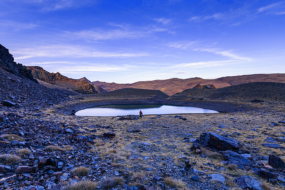 A hiker walking beside a reflective alpine lake in the high mountains of Cerro del Caballo, Sierra Nevada National Park, Andalusia, Spain.