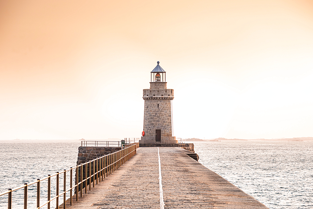 A warm sunset view of the lighthouse at Saint Peter Port’s pier, Guernsey, Channel Islands. The stone pathway stretches into the glowing horizon, where the lighthouse stands tall against a pastel sky