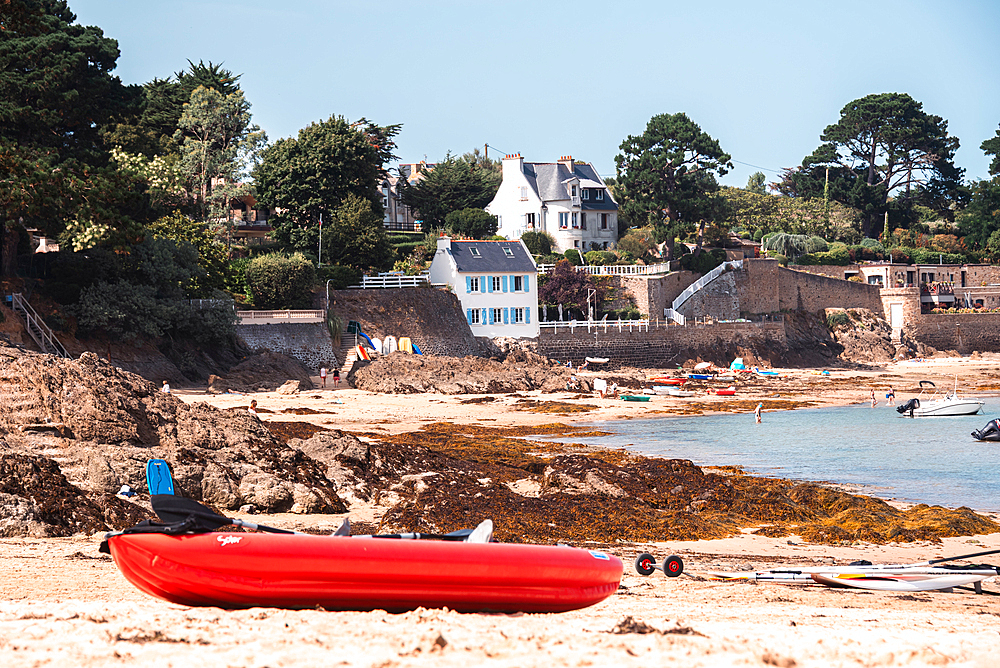 A bright red canoe rests on the rocky beach of Rotheneuf, Brittany, France, with charming coastal houses and pine trees in the background.