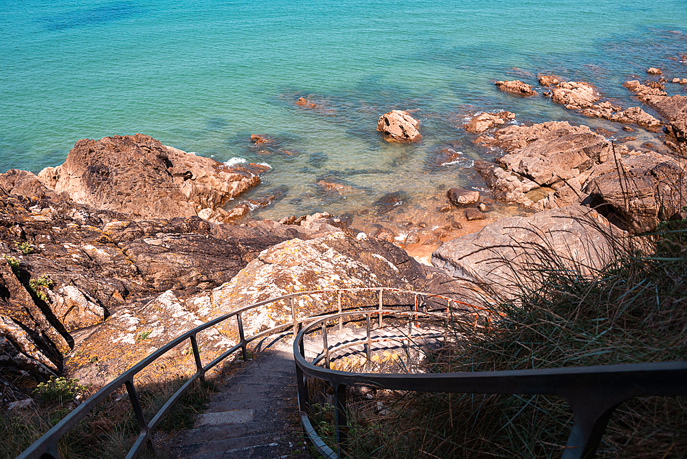 A charming stone staircase descends toward the rocky shoreline and crystal-clear waters of Brittany, France