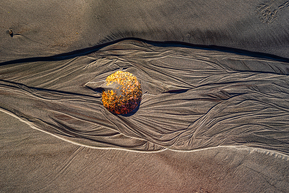 Close-up of a solitary spherical boulder resting on dark sand with rippling erosion patterns. Captures the texture natural artistry and timeless beauty of New Zealand’s rugged coastline.