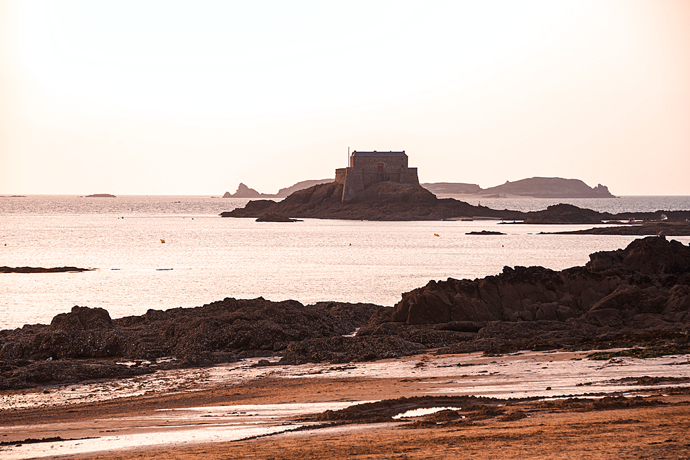 Silhouette of Grand Bé tidal island and ancient fort beyond Saint-Malo’s rocky beach with pastel sunset sky reflecting Brittany’s maritime and literary allure