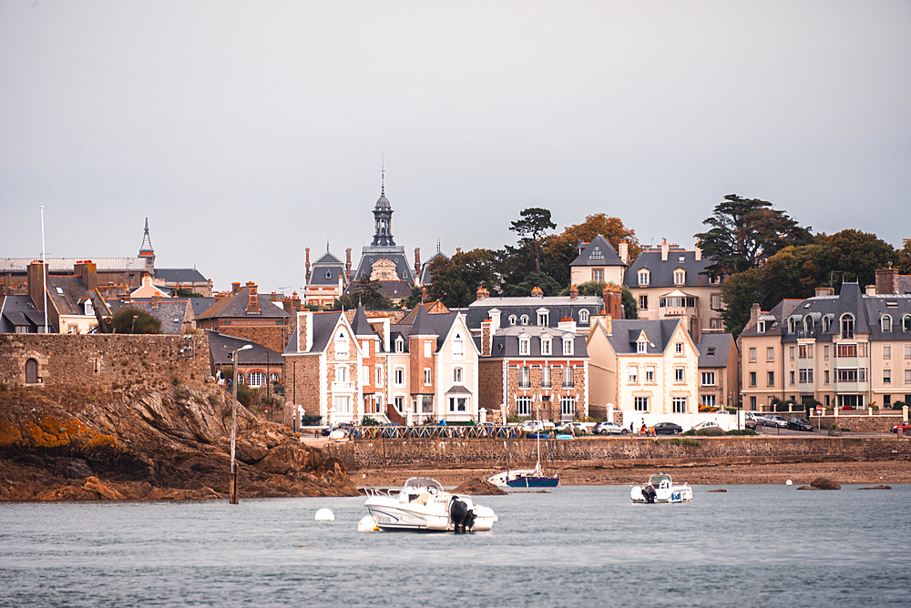 Historic stone houses and waterfront buildings line the riverbank of the Rance estuary .Le Rosais, Brittany, France. Yachts and small boats are anchored along the calm shoreline.