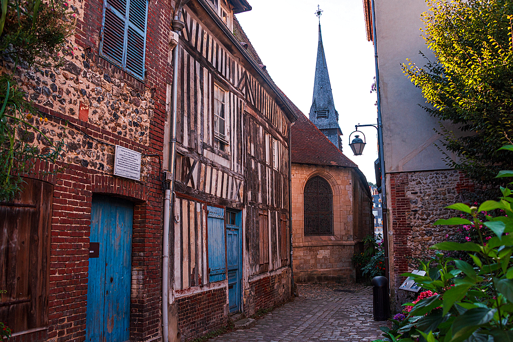 Narrow cobbled street with medieval half-timbered houses in Honfleur’s historic district, with church spire in the background.