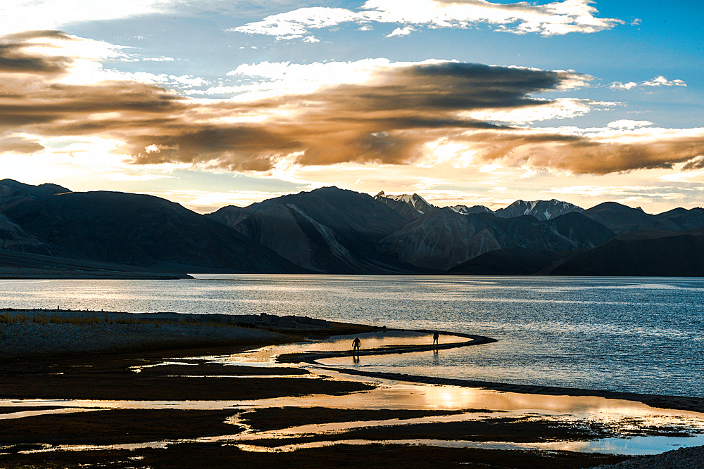 Sunset at Pan Gong lake with golden light reflection in the flat waters showing two people walking along the shore, Ladakh, northern India, Asia