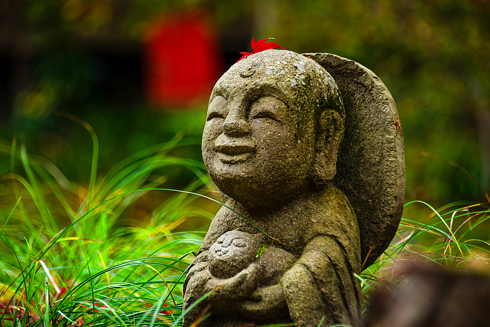 A joyful Warabe-Jizo statue cradling a child, decorated with a red maple leaf, amidst vibrant autumn grass and leaves.