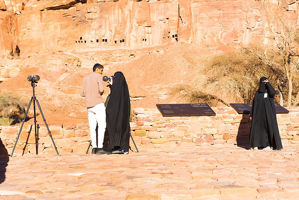 People observing, with binoculars, the tombs cut into the red rock cliffs at Archaeological site of Dadan, the ancient capital of the Dadan And Lihyan kingdoms more than 2000 years ago. AlUla, Medina Province, Saudi Arabia, Middle East