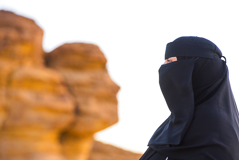 Woman wearing a Niqab posing in front of the 'Face Rock', a sandstone rock formation shaped like the human head profil, within the UNESCO World Heritage Site of Hegra, AlUla, Medina Province, Saudi Arabia, Middle East