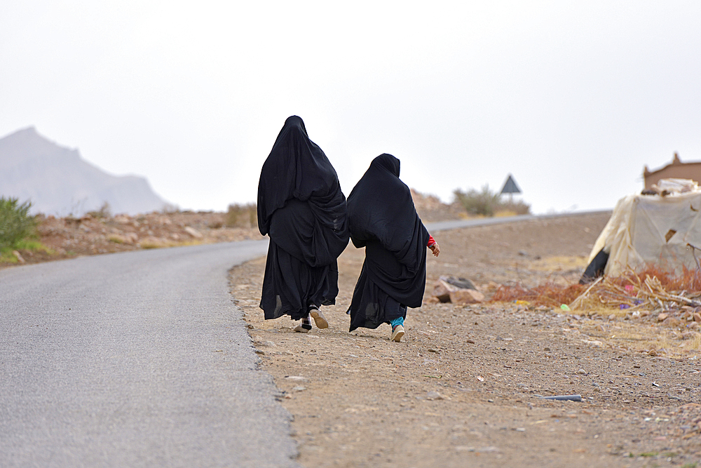 Women dressed in black abaya walking along the road, Draa River valley, Province of Zagora, Region Draa-Tafilalet, Morocco, North West Africa