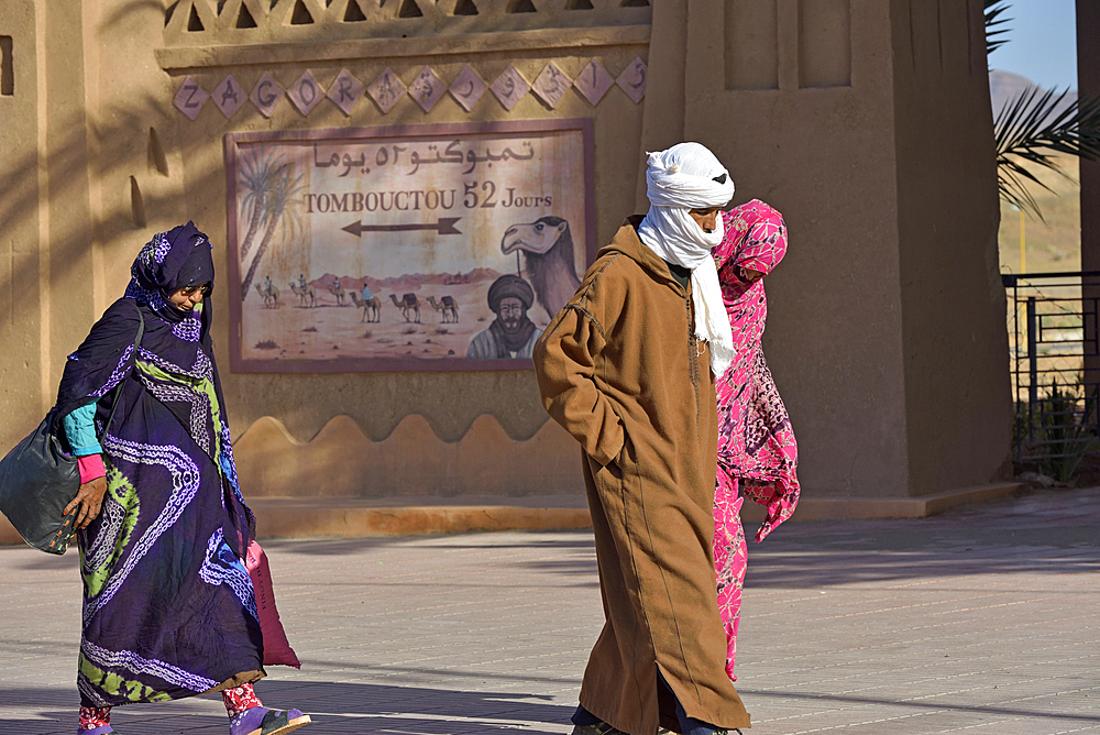 People passing by the painted sign indicating the direction of Timbuktu at 52 days of camel travel, Zagora, Draa River valley, Province of Zagora, Region Draa-Tafilalet, Morocco, North West Africa