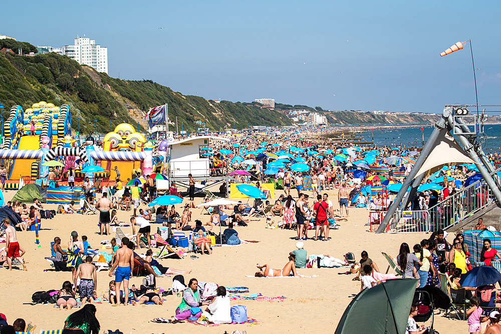 Crowded beach scene with colorful umbrellas, sunbathers, and a vibrant fairground on a sunny day in Bournemouth, Dorset, England, United Kingdom, Europe