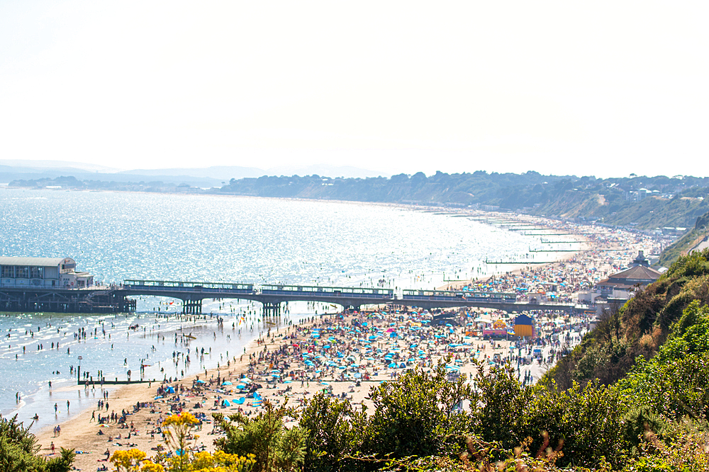 Sunny beach day with crowded sandy shore and pier, clear blue water, and lush greenery in the foreground in Bournemouth, Dorset, England, United Kingdom, Europe