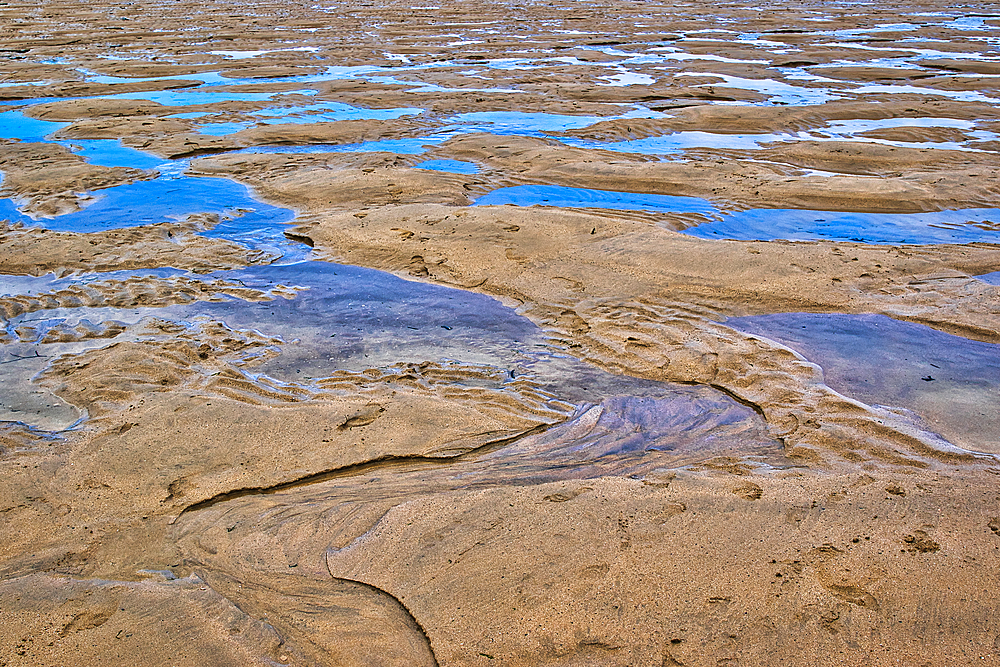 A close-up view of a sandy beach with shallow water pools. The sand features ripples and patterns created by the tide, with reflections of the sky visible in the water. The overall scene conveys a serene coastal atmosphere.