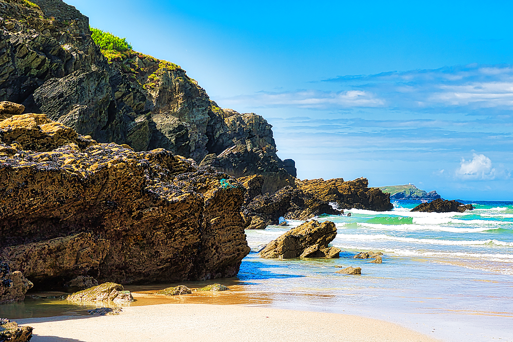 A picturesque beach scene featuring rugged cliffs and rocky formations along the shoreline. The waves gently lap at the sandy beach under a bright blue sky with scattered clouds, creating a serene coastal atmosphere.