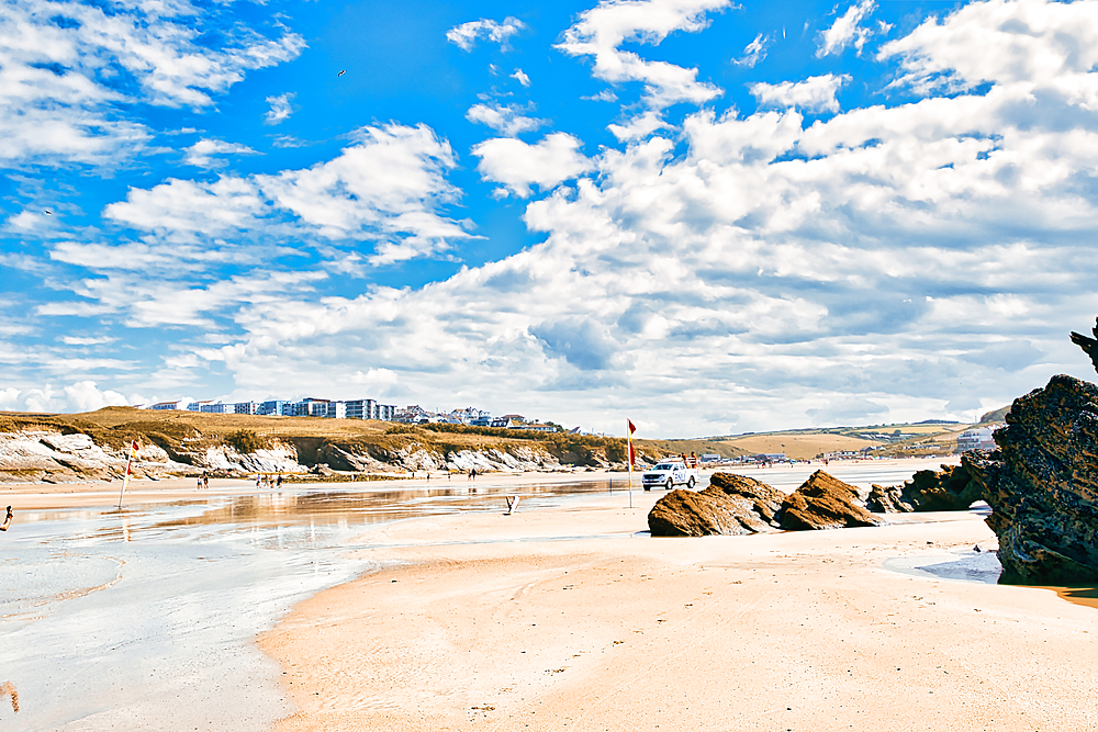 A scenic beach view featuring golden sand, gentle waves, and a clear blue sky with fluffy clouds. In the background, there are buildings on a hillside, and a few people enjoying the beach. The foreground includes rocks and a lifeguard flag.