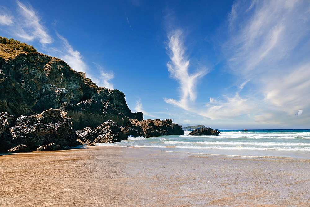 A scenic beach view featuring golden sand, rocky cliffs, and a vibrant blue sky with wispy clouds. The waves gently lap at the shore, creating a tranquil atmosphere. Ideal for nature and travel themes.