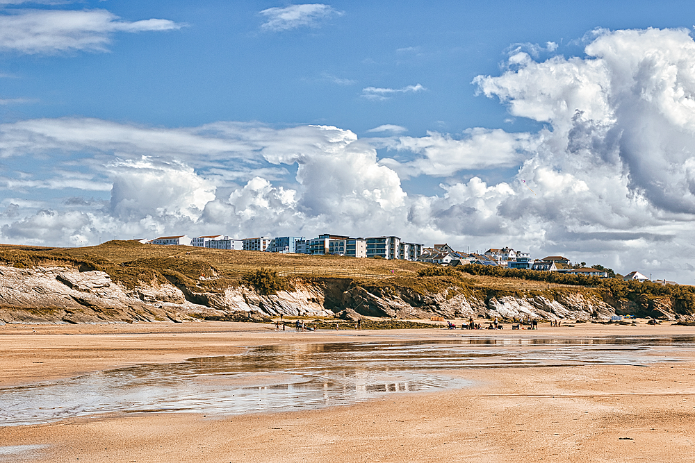 A scenic beach view featuring a sandy shore with gentle waves, rocky cliffs, and a backdrop of white clouds. In the distance, modern buildings are situated on a hill, contrasting with the natural landscape. The sky is bright and clear, enhancing the tranq