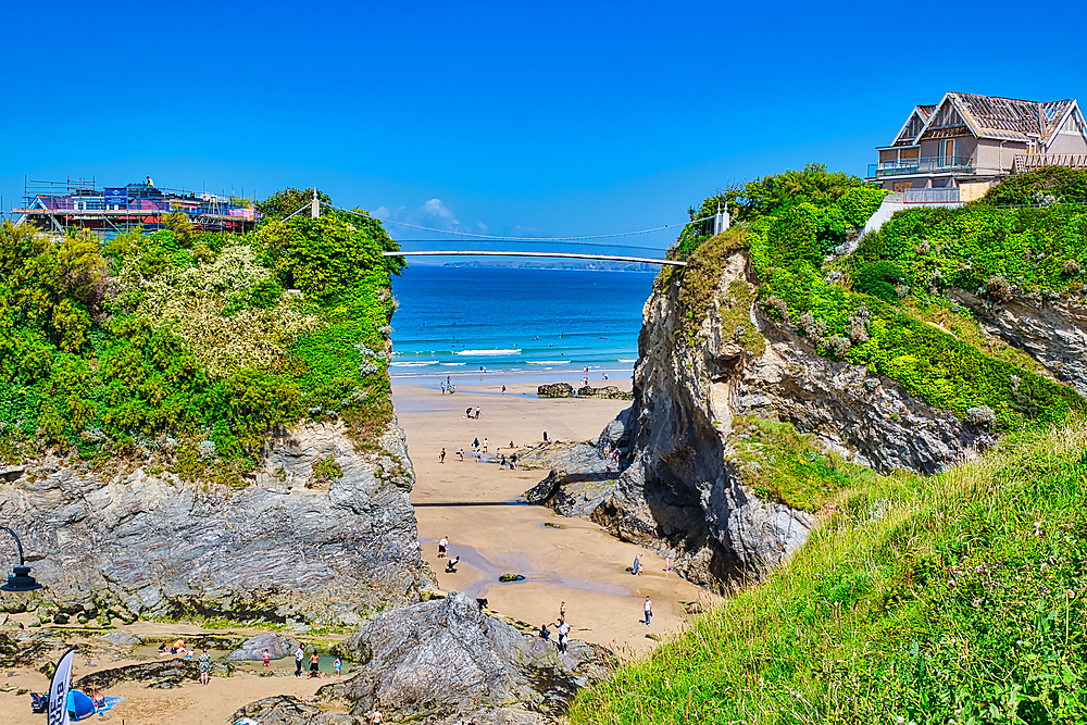 A scenic beach view featuring cliffs on either side, with people walking on the sandy shore. The ocean is calm and blue, and a bridge can be seen in the distance. Lush greenery adorns the cliffs, and a clear blue sky completes the picturesque scene.