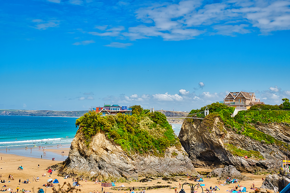 A scenic beach view featuring rocky cliffs, sandy shores, and people enjoying the sun. The sky is bright blue with scattered clouds, and there are houses perched on the cliffs overlooking the ocean.