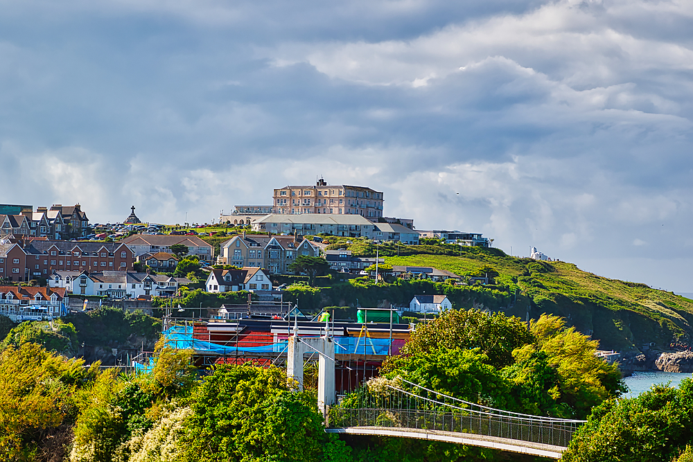 A scenic view of a coastal town with a hotel on a hill, surrounded by lush greenery and colorful boats in the harbor. The sky is partly cloudy, creating a dramatic backdrop.