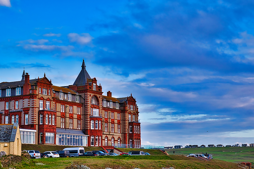 A historic red brick hotel building with a turret, set against a blue sky with scattered clouds. The hotel is situated on a grassy hill, with several parked cars in the foreground and a distant view of the coastline.