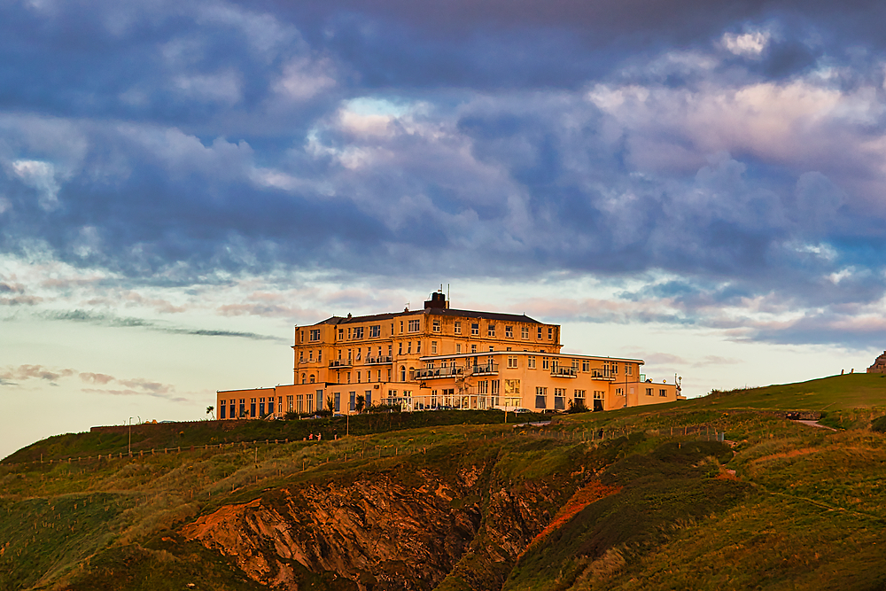 A large, elegant hotel perched on a cliff overlooking the ocean. The building features multiple stories with large windows and a balcony, surrounded by lush green grass and rocky terrain. The sky is filled with dramatic clouds, creating a picturesque suns