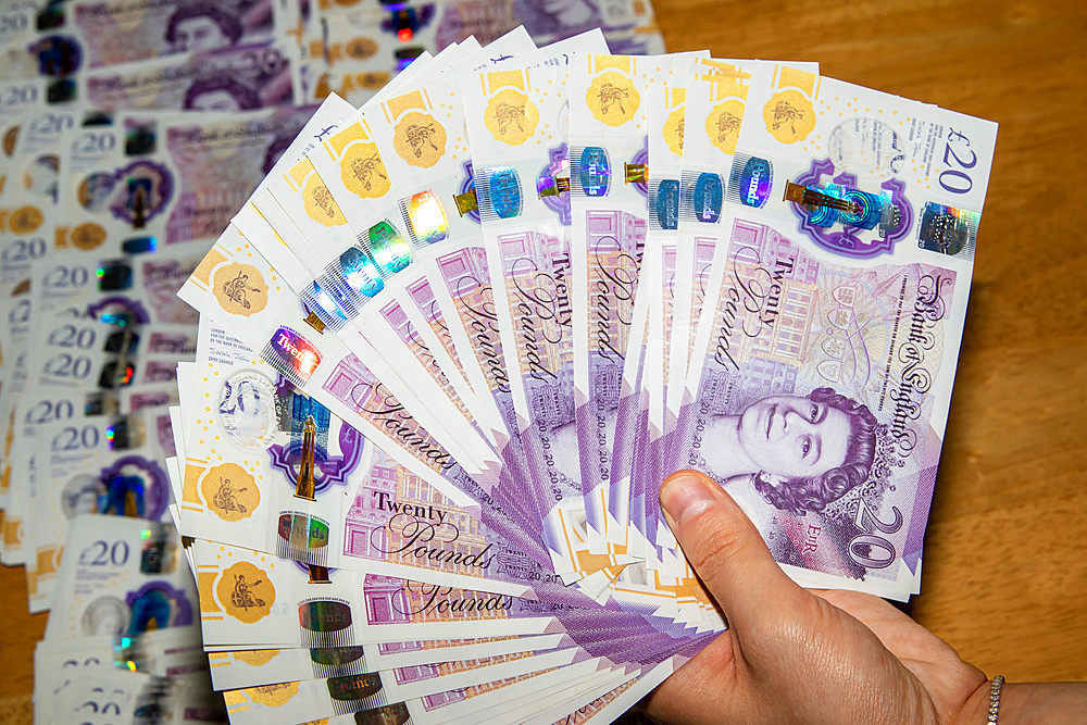 A hand holding a fan of twenty-pound banknotes, showcasing the vibrant colors and holographic features of the currency. In the background, additional banknotes are spread out on a wooden surface.