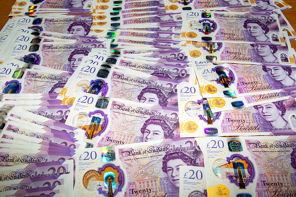 A large spread of British twenty-pound banknotes arranged on a flat surface. The notes feature intricate designs, holographic elements, and the portrait of Queen Elizabeth II. The colors are predominantly purple with gold accents.