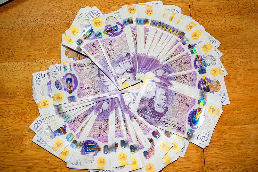 A circular arrangement of British twenty-pound notes displayed on a wooden surface. The notes are fanned out, showcasing their purple color and intricate designs, with the Queen's portrait visible. The background is a simple wooden table.