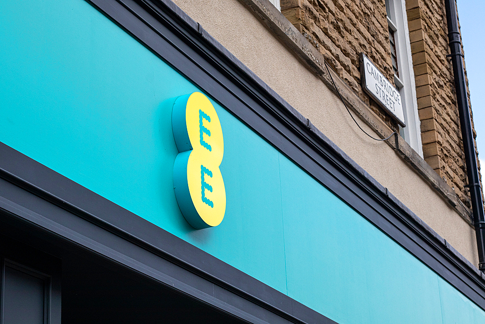 A close-up of the EE logo on a storefront, featuring a bright yellow logo against a teal background. The logo consists of two overlapping circles with the letters 'E' prominently displayed. The building has a stone facade and a sign above that reads 'CHUR