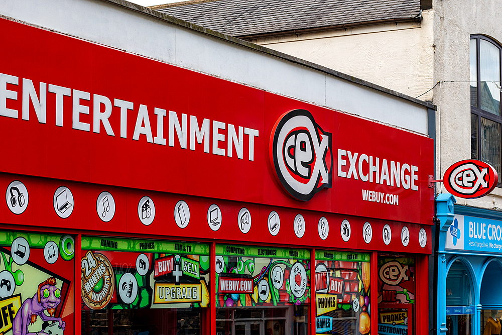 A vibrant storefront of a second-hand entertainment exchange shop, featuring bright red signage with the logo 'Cex'. The shop displays various colorful graphics related to entertainment, including music notes and gaming icons. Nearby, a blue shop sign rea