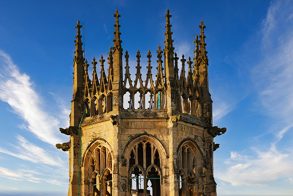 A stone tower with pointed spires against a bright blue sky with white clouds. The tower is detailed with intricate stonework and gargoyles.