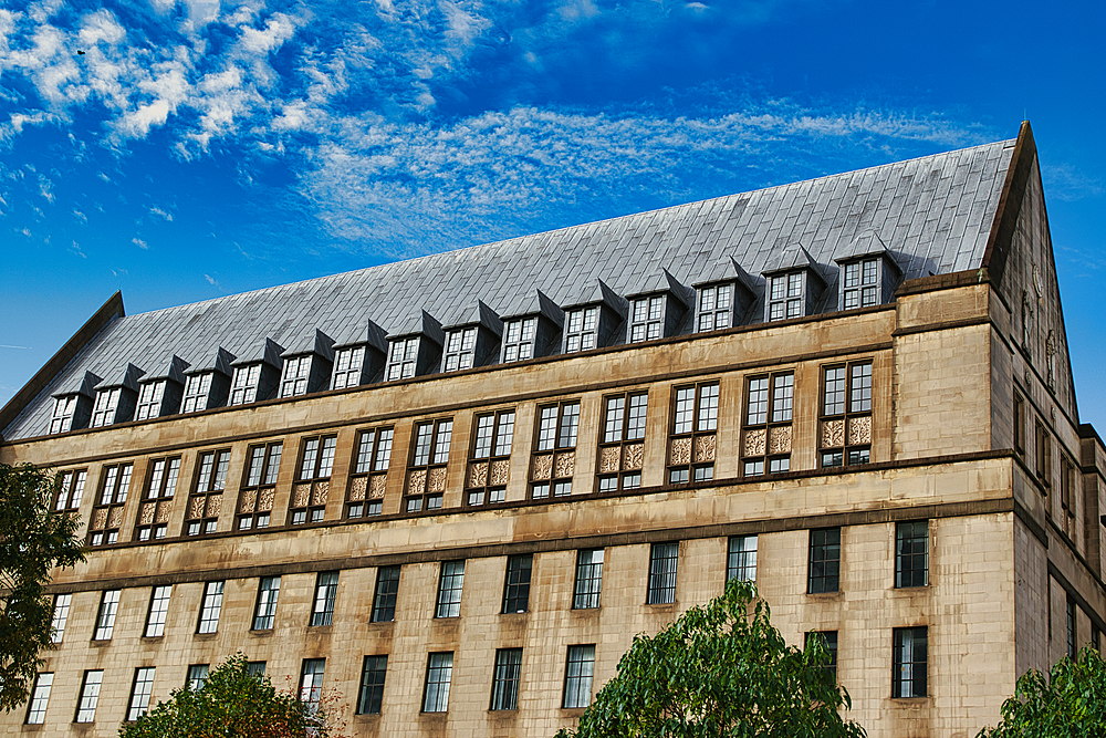 A low-angle view of a large, beige building with a dark gray roof and many windows. The building is set against a blue sky with white clouds in Manchester, UK.